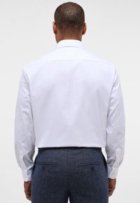 Camicia bianca a maniche lunghe con colletto classico, tessuto morbido e polsini con bottoni, abbinata a pantaloni blu scuro con texture e tasche posteriori.