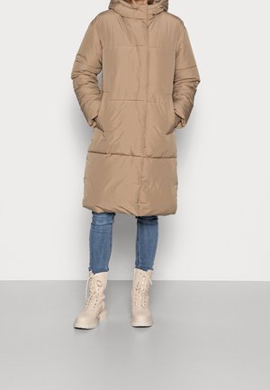 Personne portant un manteau matelassé beige à capuche jusqu'aux genoux, un jean skinny bleu et des bottines lacées beige clair, debout les mains dans les poches.