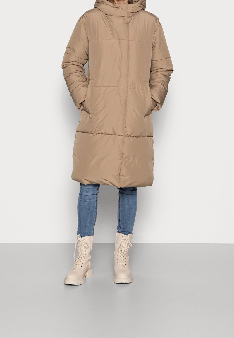 Personne portant un manteau matelassé beige à capuche jusqu'aux genoux, un jean skinny bleu et des bottines lacées beige clair, debout les mains dans les poches.