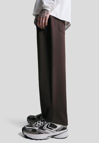 Bruine, loszittende broek van gladde stof, gecombineerd met zwart-zilveren sneakers met mesh details en lichte zolen.