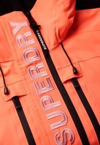 Jasnopomarańczowa kurtka z zamkiem błyskawicznym z przodu, teksturowaną tkaniną i haftowanym napisem "SUPERDRY" na pionowym pasku, z czarnymi detalami przy zamku.