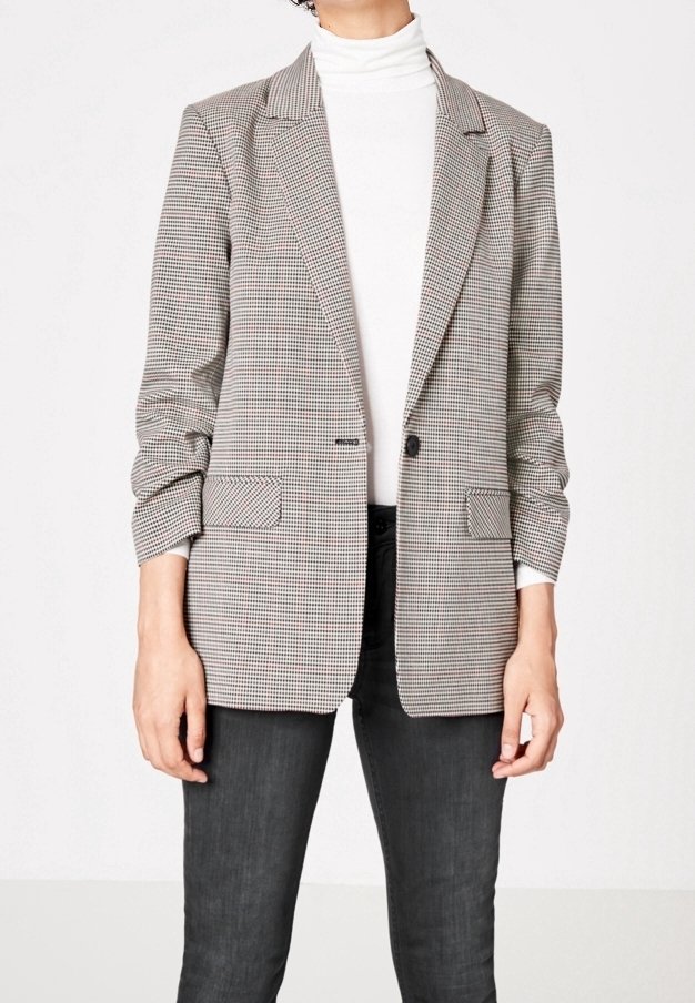 Personne portant un blazer pied-de-poule beige et noir sur un col roulé blanc, associé à un pantalon ajusté gris foncé.