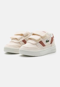 Sneakers Lacoste in tela beige chiaro con due strappi in velcro, accentuate da una striscia marrone e un logo verde, caratterizzate da una superficie testurizzata.