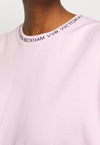 Světle růžové bavlněné tričko s kulatým výstřihem, které má černý potisk loga kolem límce. Hladká textura, jednoduchý design.