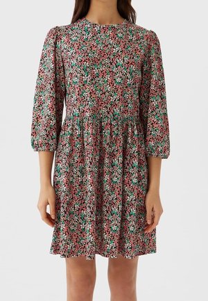 Vrouw die een knielange bloemenjurk draagt met driekwart mouwen en een hoge halslijn op een lichtgrijze achtergrond.