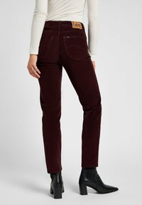 Maroon corduroy pants with a straight fit, featuring rear pockets and a brown leather Lee logo patch at the waistband. Worn with black heeled boots.