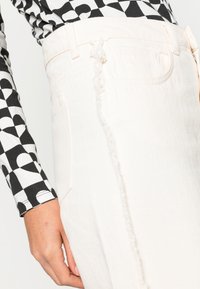 Calças de ganga brancas com detalhes de costura desfiada. O tecido tem um acabamento texturizado. O modelo usa uma blusa de manga longa com padrão preto e branco.