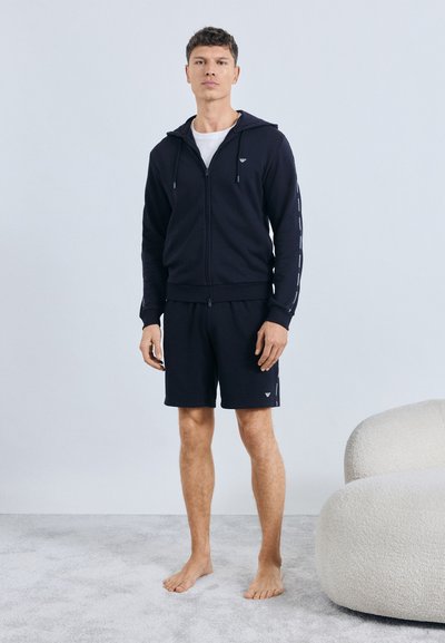 Tengerészkék kapucnis pulóver cipzárral és húzózsinórokkal, logócsíkokkal és letisztult dizájnnal, a hozzáillő tengerészkék rövidnadrággal párosítva.