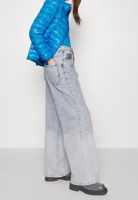 Světlé modré široké džíny s vybledlou texturou, vysokým pasem a předními kapsami, doplněné těsně padnoucí modrou prošívanou bundou a šedými botami.