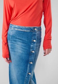 Jupe en denim à boutons sur le devant avec une taille haute, couleur bleu clair, coutures visibles et cinq boutons blancs à l'avant. Haut orange à manches longues.