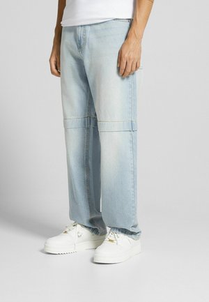 Homme portant un jean denim bleu clair ample et des baskets blanches, debout devant un fond gris uni.