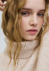 Krém színű, magas nyakú pulóver, texturált kötött mintával. Hosszú, hullámos haj részben eltakarja a pulóver nyakát.