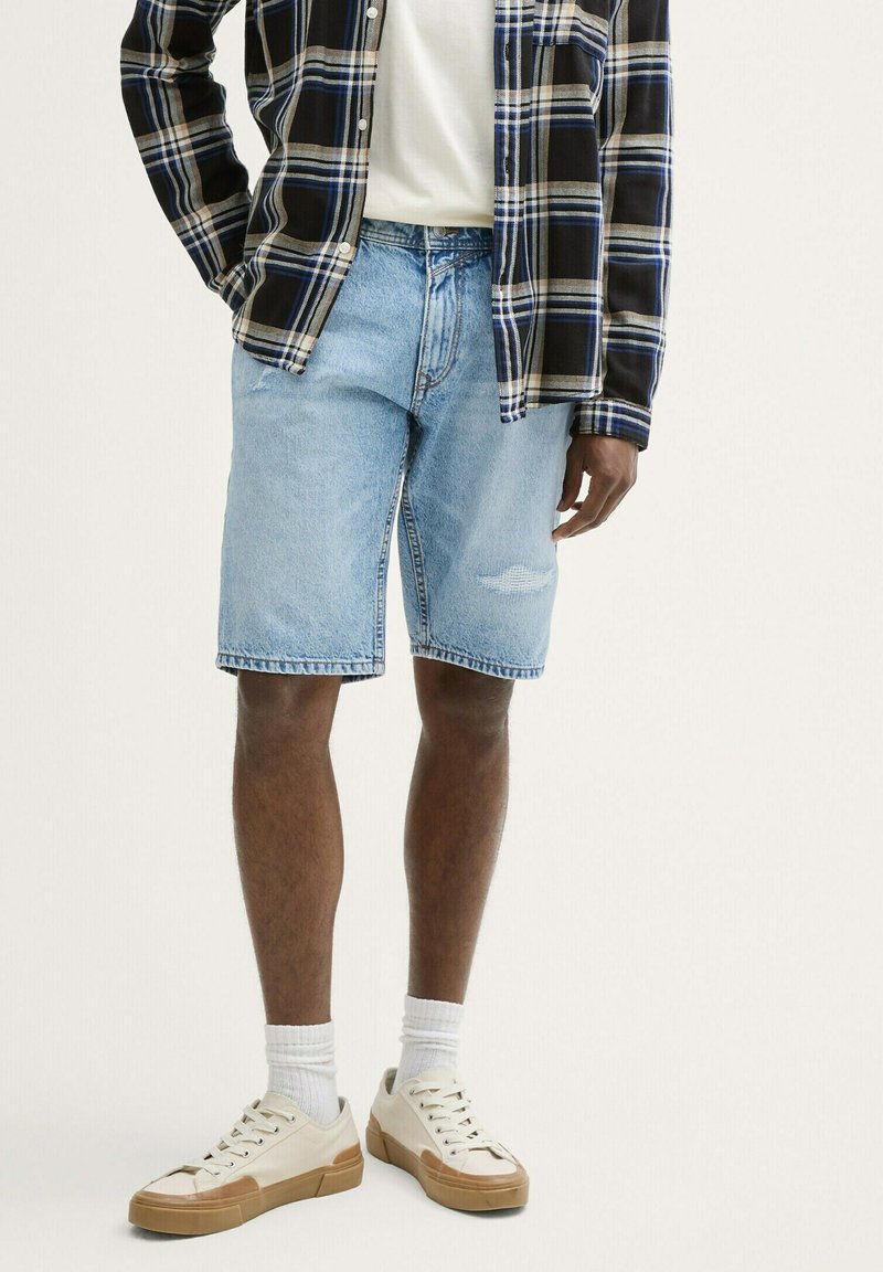 Pantalones cortos de mezclilla de color azul claro con un corte relajado, que presentan un patrón desgastado. Combinados con una camisa de cuadros en negro y azul y zapatillas blancas.