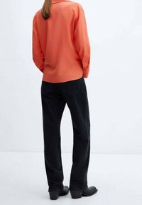 Blusa arancione a maniche lunghe con colletto, abbinata a jeans neri a gamba larga. Tessuto liscio, con vista posteriore che mostra tasche e calzature nere.