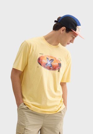 Geel katoenen t-shirt met een grafische print met de tekst "PRESS START 1UP" in rood en zwart. Gepaard met kaki shorts en een blauwe pet.