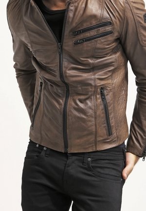 Homme portant une veste en cuir marron ajustée avec plusieurs fermetures éclair noires et un pantalon noir, debout avec une main dans la poche.