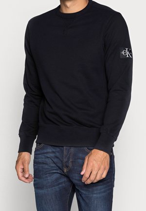 Mand iført en sort langærmet sweatshirt med et "CK Calvin Klein" mærke på venstre ærme og blå jeans.