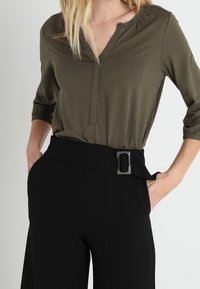 Vrouw in een olijfgroene V-hals top met driekwart mouwen en hoge zwarte broek met een brede riem en een rechthoekige gesp.