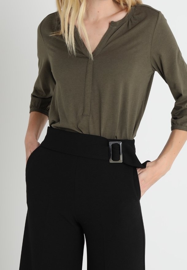 Vrouw in een olijfgroene V-hals top met driekwart mouwen en hoge zwarte broek met een brede riem en een rechthoekige gesp.