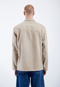 Chaqueta de algodón beige con un cuello con solapa y mangas largas, con acentos de costura, vista desde la parte trasera, acompañada de pantalones de mezclilla azul casuales.