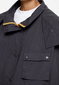 Veste d'extérieur noire avec un col montant, un détail de fermeture éclair jaune et une poche à bouton-pression. Texture de tissu lisse avec des éléments de design structurés.