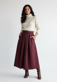 Bordowa plisowana spódnica z pętlą na pasek i okrągłą klamrą, zestawiona z kremowym prążkowanym golfem. Brązowe skórzane botki za kostkę.