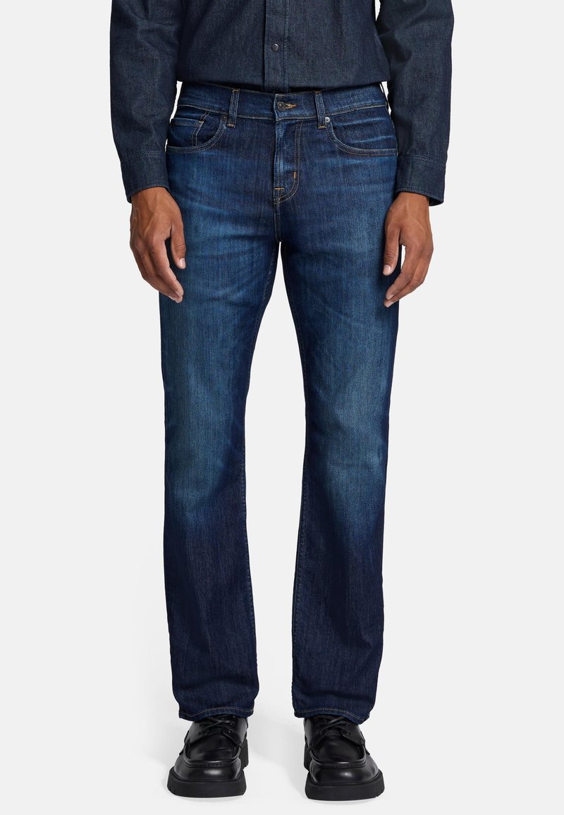 Jeans in denim blu scuro con una linea dritta, caratterizzati da cinque tasche e cuciture gialle. Indossati con scarpe nere e una camicia button-up scura.