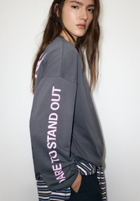 Szürke hosszú ujjú pulóver rózsaszín "MERJ KÜLÖNLEGES LENNİ" felirattal a bal ujján. Csíkos szegéllyel az alján. Simább textúra.