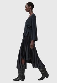 Kvinde går iført sort indpakningsstrik, plisseret midinederdel og sorte broderede cowboystøvler på en ensfarvet grå baggrund.