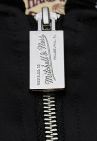 Silberner Metall-Reißverschluss-Zipper mit eingraviertem "Mitchell & Ness", befestigt an schwarzem Stoff mit sichtbaren Zähnen und Nähten.