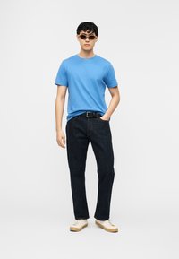 Νέος άνδρας που φοράει καφέ γυαλιά ηλίου, μπλε κοντομάνικη μπλούζα, σκούρο τζιν με μαύρη ζώνη, λευκά αθλητικά παπούτσια, στέκεται με το ένα χέρι στην τσέπη.