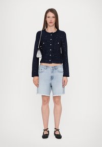 Marine teksturert cardigan med dekorative knapper, lyseblå denimshorts med frynset kant, og svarte leppestifter med prikkete mønster.