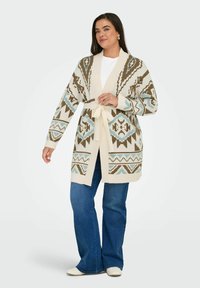 Ženska v modrih kavbojkah in belih čevljih, s bež, modro in rjavo vzorčasto kardiganom s pasom čez belo majico, stoji z rahlim nasmehom.