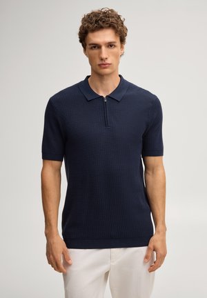 Junger Mann mit lockigem Haar, der ein marineblaues kurzärmeliges Strick-Poloshirt mit Frontreißverschluss und weiße Hose trägt, steht vor einfachem Hintergrund.