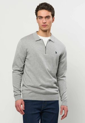 Młody mężczyzna w jasnoszarej, długorękawowej bluzie polo z suwakiem do ćwiartki i ciemnoniebieskich dżinsach stoi na jednolitym tle.