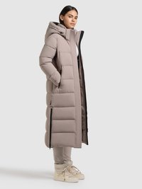 Lange gewatteerde jas in licht beige met een capuchon, zijzakken met rits en een doorgestikte textuur. Gedragen over beige broek en lichtgekleurde sneakers.