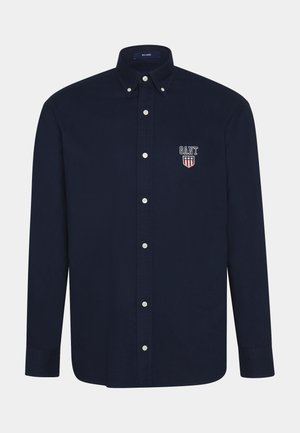 Camisa de algodón azul marino abotonada con cuello, botones blancos y logo bordado de GANT con diseño de escudo en el lado izquierdo del pecho.