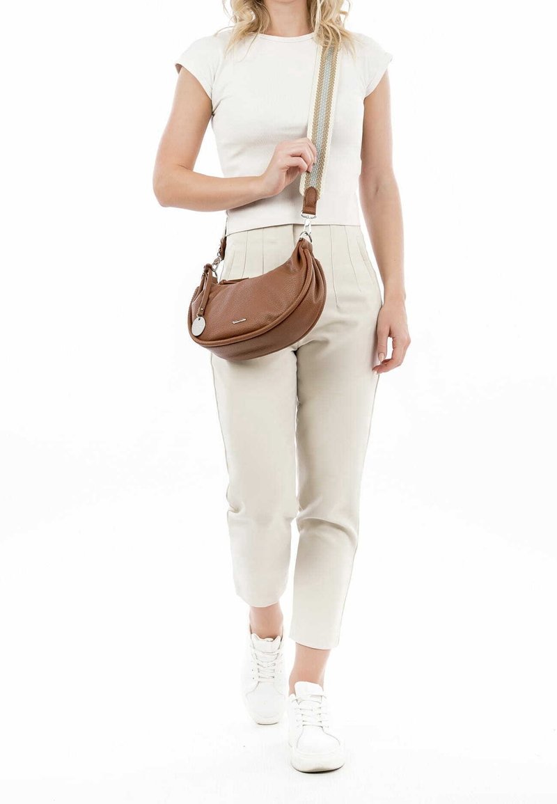 Femme portant un pantalon beige et un haut blanc, avec un sac en cuir marron porté en bandoulière avec une large sangle rayée et des baskets blanches.