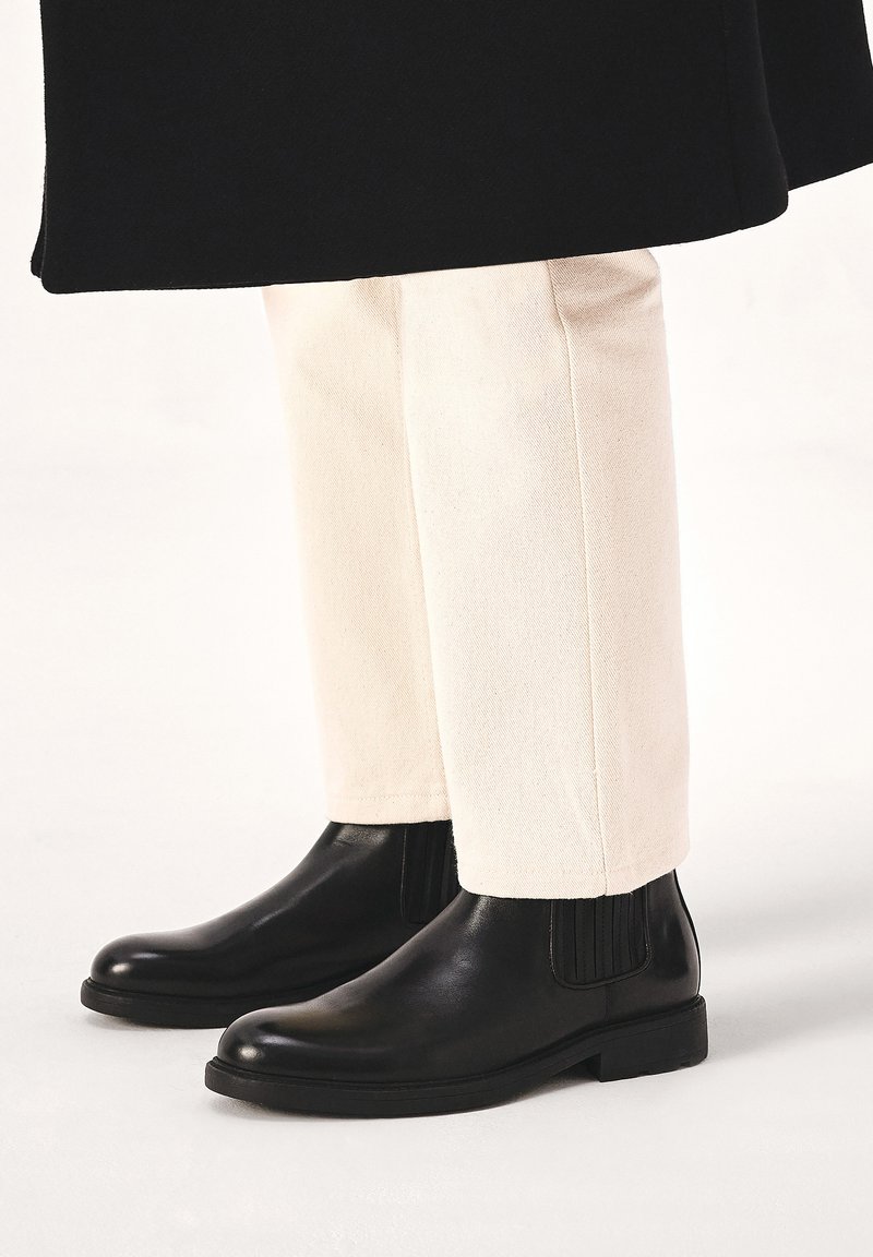 Botas de cuero negro hasta el tobillo con paneles elásticos a los lados, combinadas con pantalones de corte tapered en color crema. Textura suave y diseño clásico.