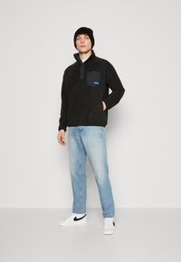 Pullover in pile nero con colletto e chiusura a bottone, caratterizzato da una tasca scura e un logo blu. Abbinato a jeans azzurri chiari e sneakers bianche.