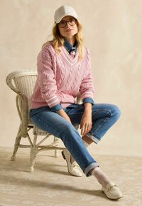 Hele roosa soonikkoonatud sviiter valgete triipudega, teksased, valged loafers'id, muster sokid ja beež müts, istub viinapuu toolil.