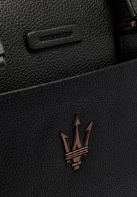 Sac en cuir noir texturé avec un logo trident en bronze et une petite étiquette embossée portant l'inscription "Piquadro". Design simple et rectangulaire.