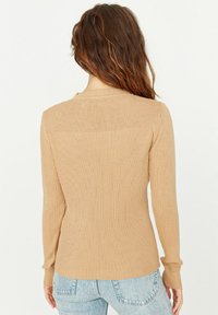 Beiges, geripptes Strick-Oberteil mit langem Arm, rundem Ausschnitt, figurbetonter Silhouette und strukturierten Details auf der Rückseite. Kombiniert mit hellblauen Jeans.