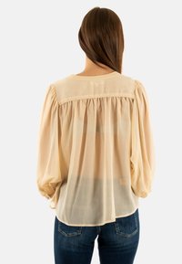 Lichtbeige blouse met pofmouwen, doorzichtige stof, geplooide achterkant en ronde halslijn, gecombineerd met donkerblauwe jeans.