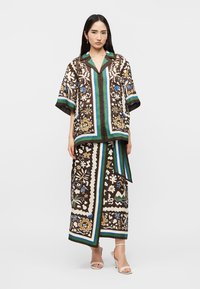 Kvinne iført brunmønstret skjorte med knapper og matchende langt skjørt med hvite høye hæler, foran hvit bakgrunn.