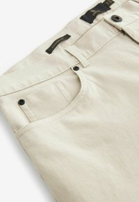 Pantalons droit texturés beiges avec deux accents de boutons noirs sur la ceinture et une poche angulaire. Étiquettes visibles à l'intérieur de la ceinture.