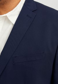 Navyblazer aus glattem Stoff, mit Revers und einer Brusttasche. Das Hemd darunter ist weiß mit einer strukturierten Oberfläche.