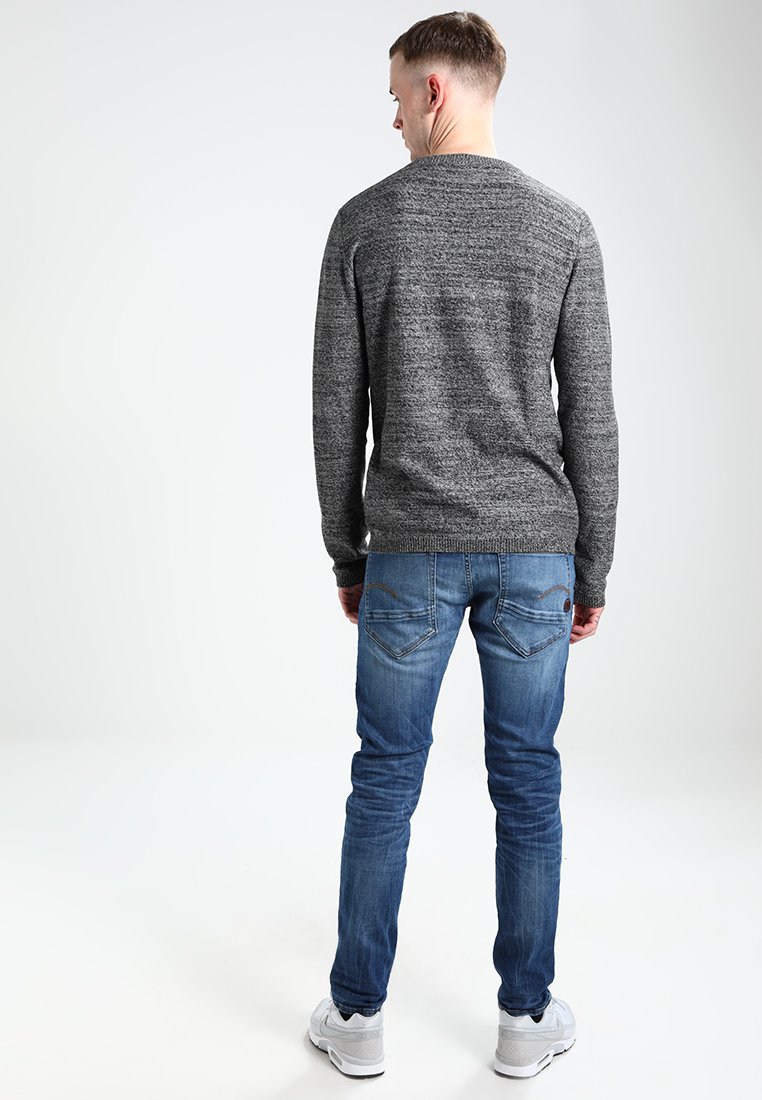 Un pull en tricot gris avec un motif texturé, porté avec un jean bleu. La tenue comprend des baskets blanches, affichant un look décontracté vu de l'arrière.