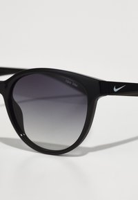 Melnas Nike Max saulesbrilles ar gradienta lēcām, apaļu rāmī un mazu sudraba Nike logotipu uz roka. Lēcas nodrošina UV aizsardzību.