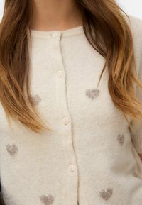 Button-up vest in crèmekleur, met hartpatronen in lichtbruin. Gemaakt van zacht materiaal met een gezellige textuur. Ronde halslijn en korte mouwen.